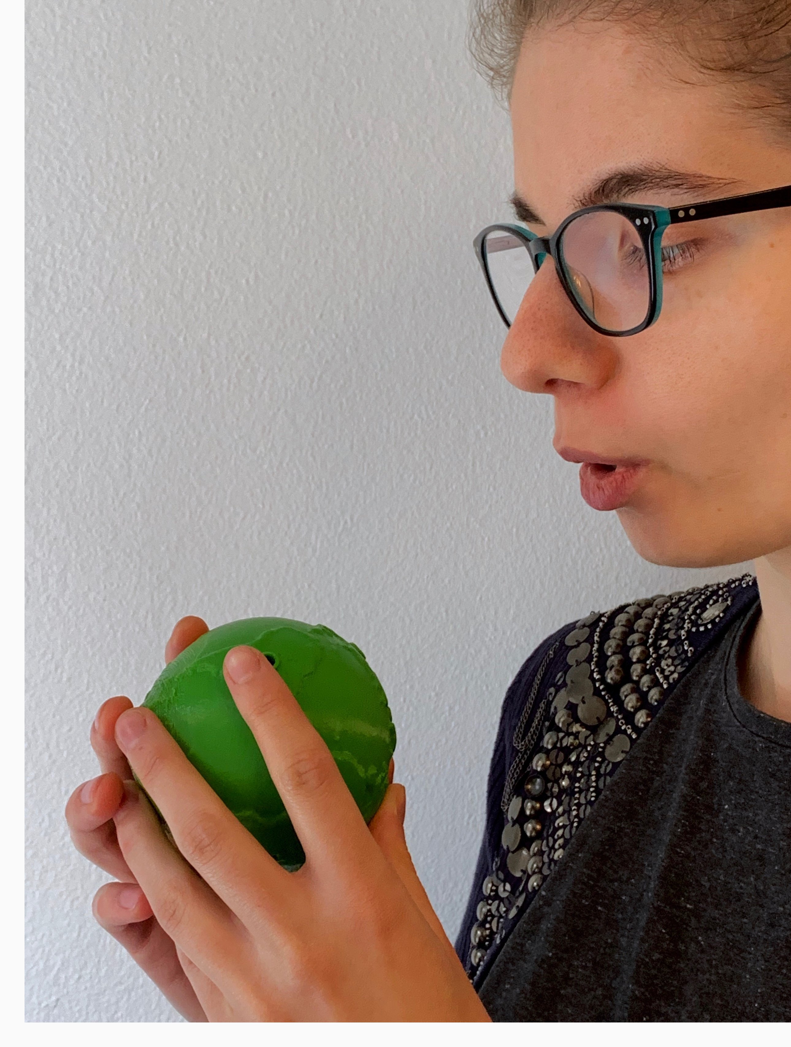 A person with glasses gently holds a green ball, blowing on it. The background is a plain white wall.