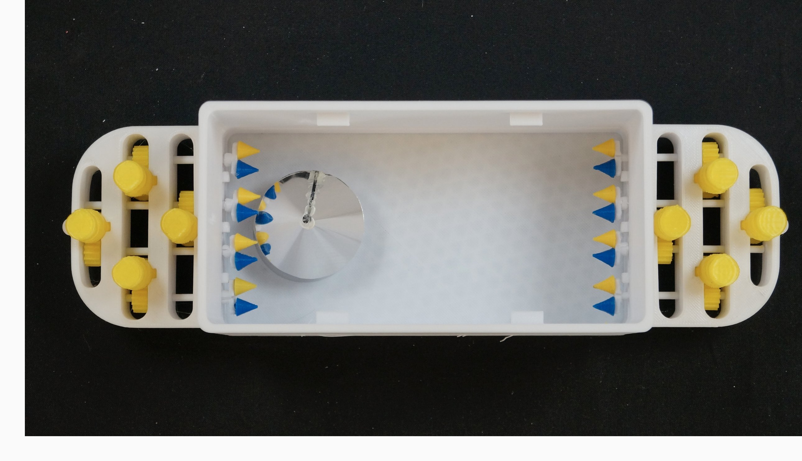 A white rectangular device with yellow knobs on both sides, featuring blue and yellow triangular spikes inside, against a black background. A silver circular disc is also visible inside.