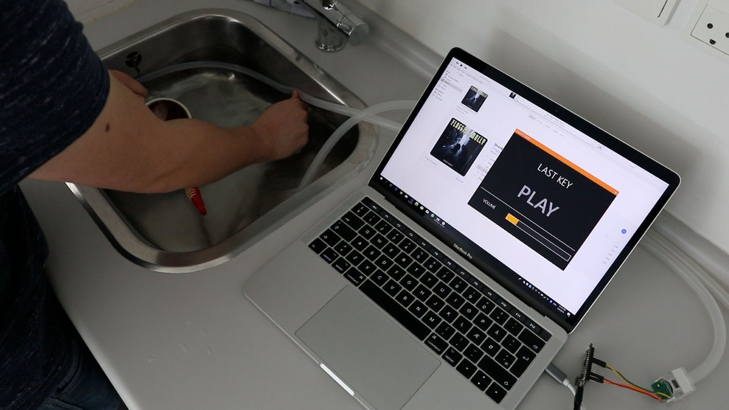 A person presses on a hose in a kitchen sink while a MacBook Pro on the counter
displays a “Music Player” screen with volume controls