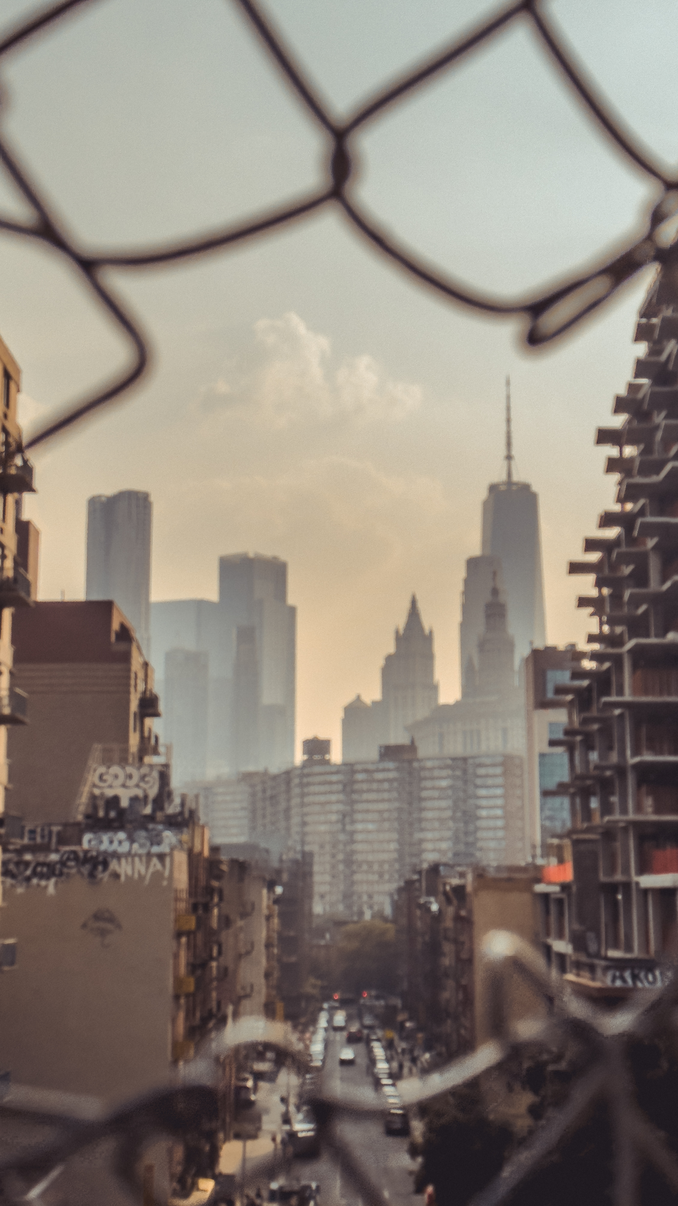 A hazy cityscape view with tall skyscrapers and apartment buildings seen
through the blurred diamond pattern of a chain-link fence, with a muted sunset
sky.