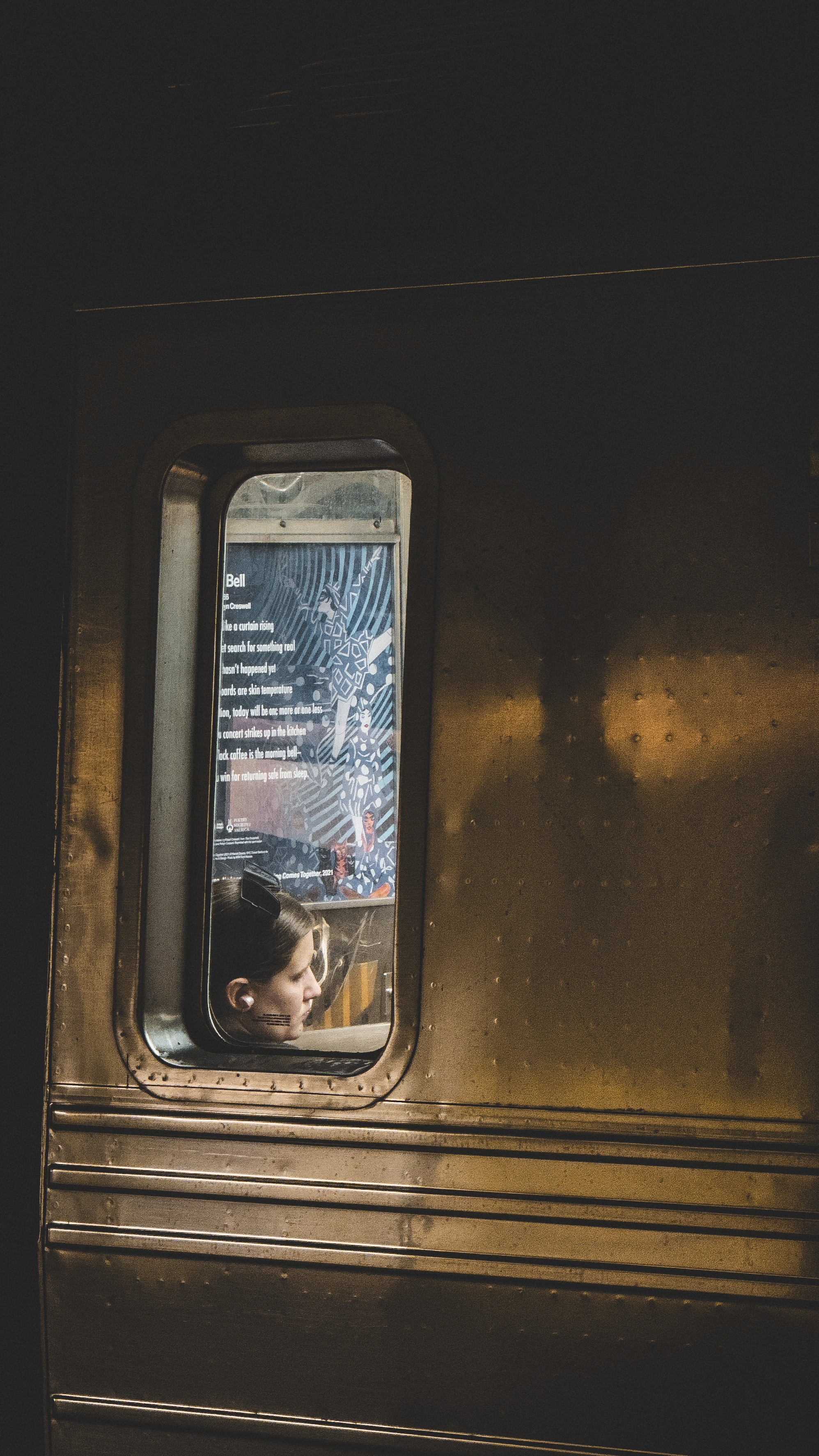 A woman with headphones, seen through the window of a dimly-lit subway train,
with a poem and abstract art posted on the wall behind
her.