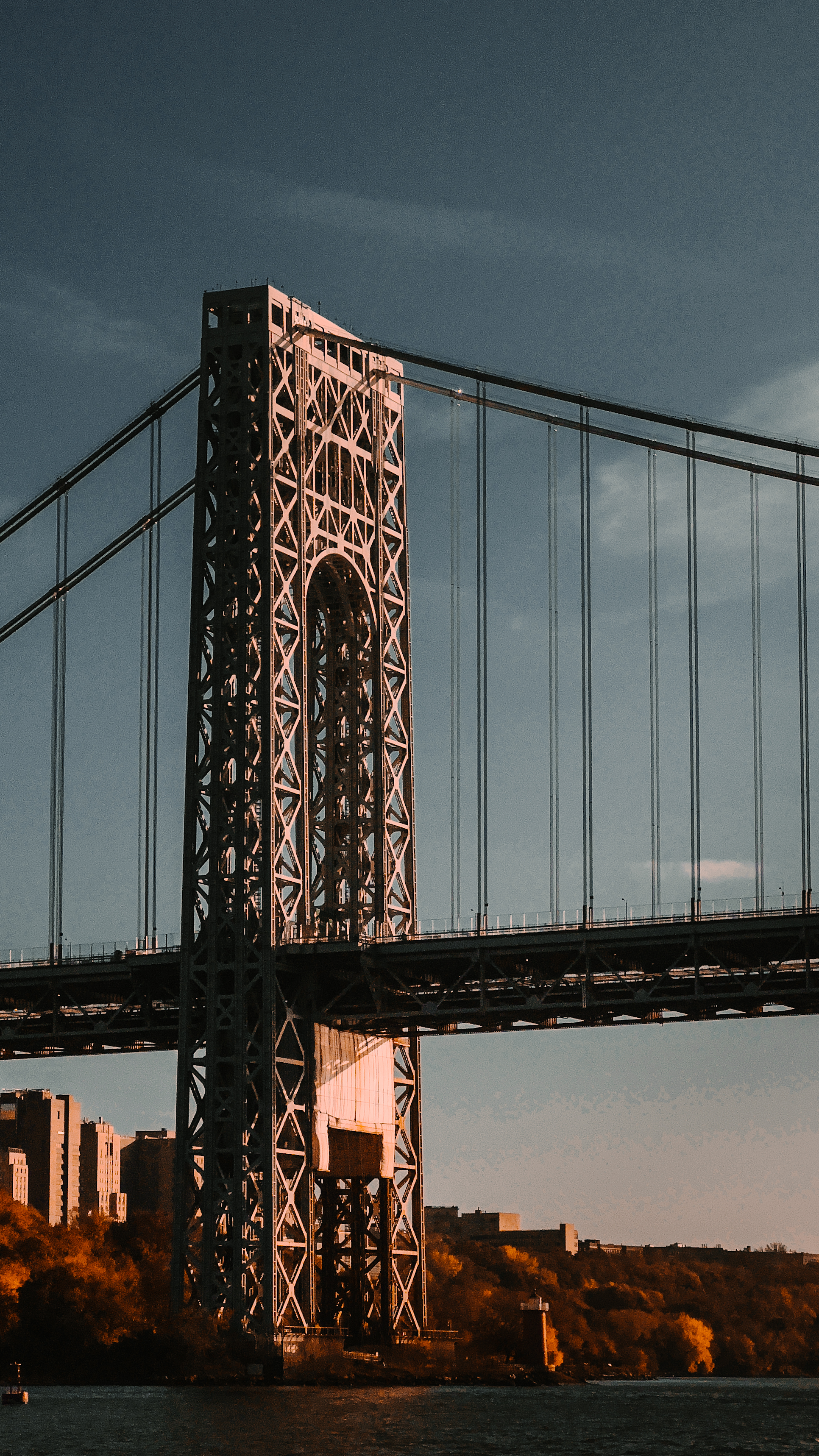 The George Washington Bridge’s steel tower and suspension cables are
dramatically lit by evening sunlight, spanning across a river with city
buildings and trees in the background.