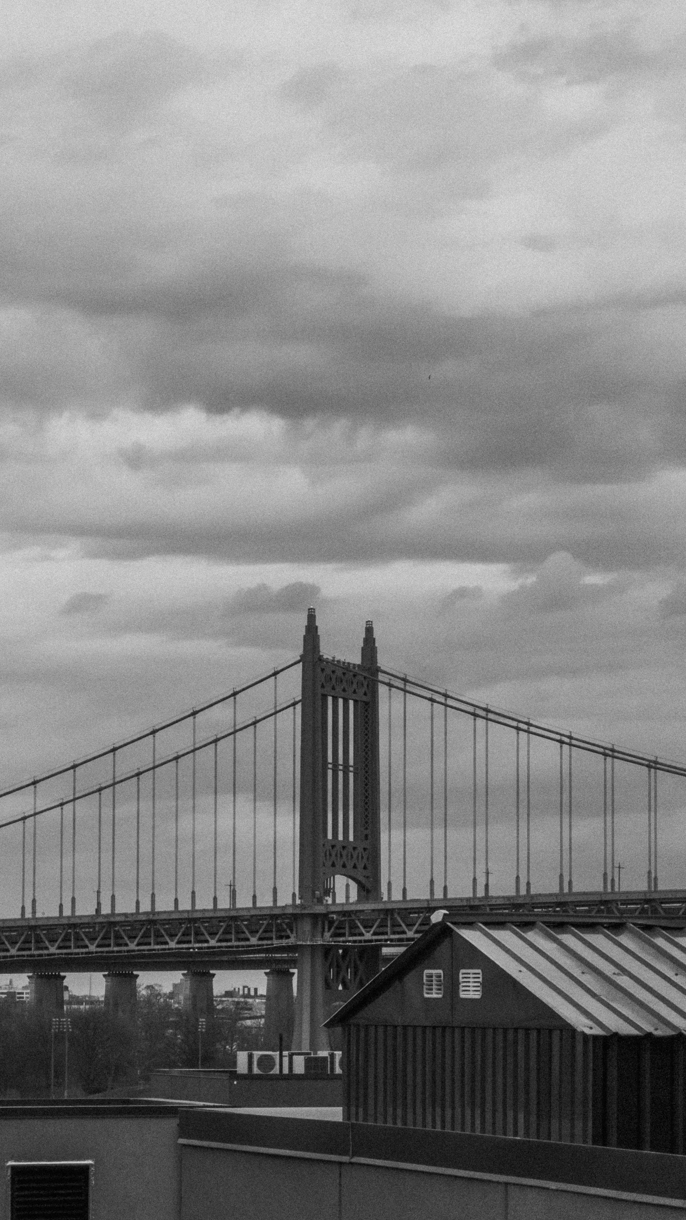 A black and white image of a suspension bridge spanning across the frame, with
dark clouds in the sky and industrial rooftops in the
foreground.