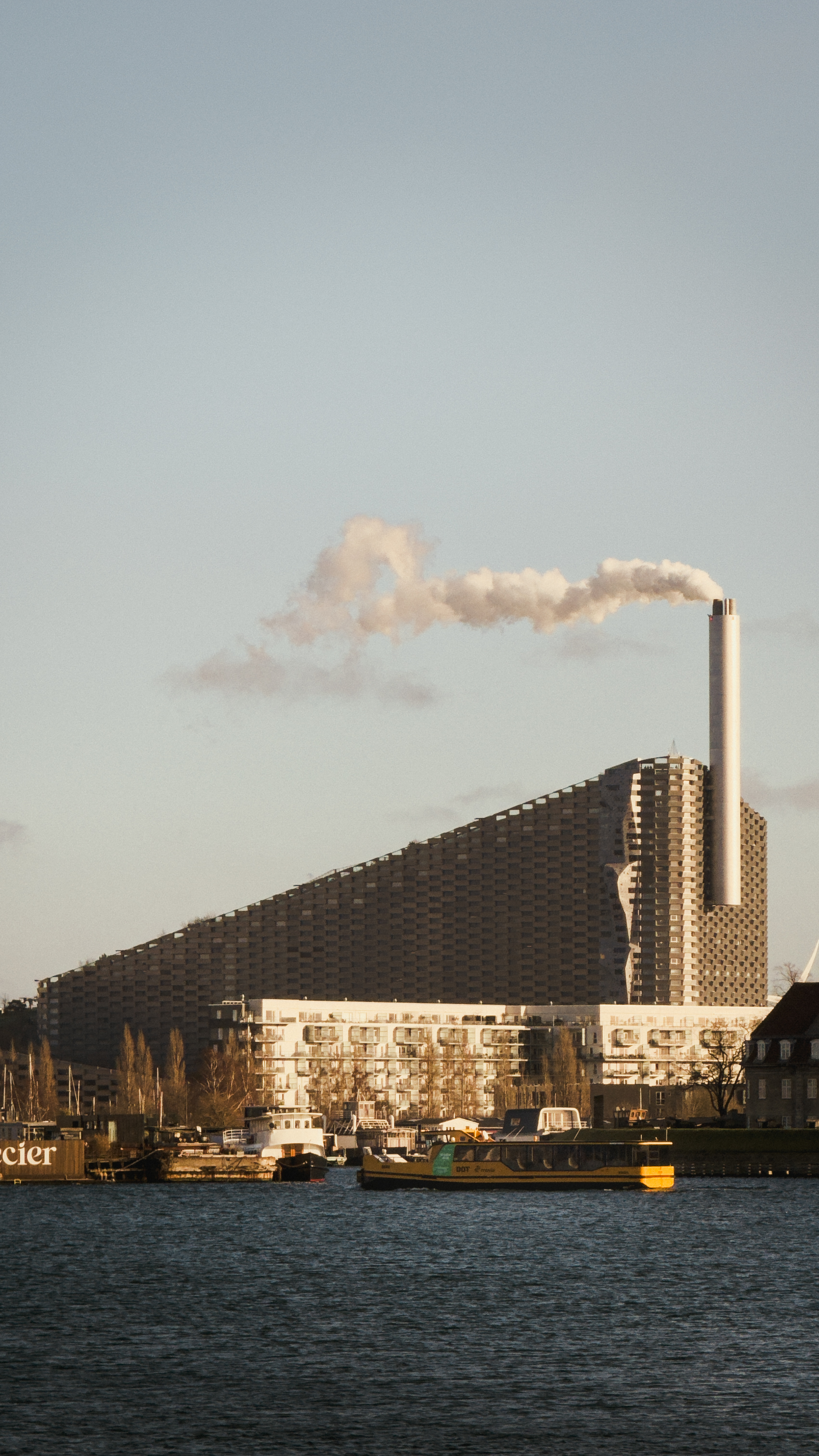 A modern waste-to-energy plant with a unique sloped roof and a tall smokestack
emitting a cloud of steam, seen from across a harbor with boats and buildings in
the foreground.