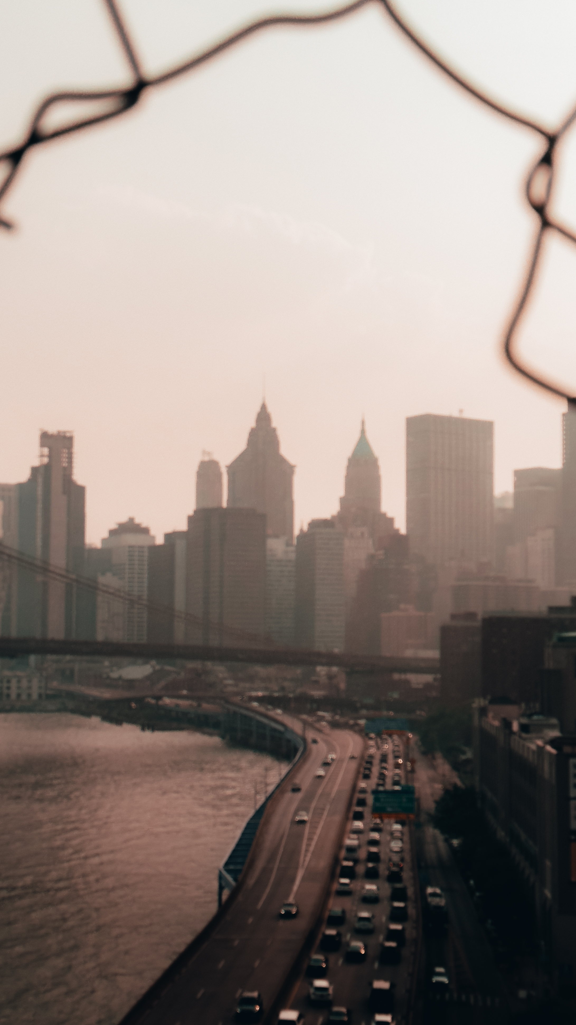 A hazy cityscape view of New York City’s skyline through a chain-link fence,
with a highway curving along the river and tall skyscrapers in the
background.