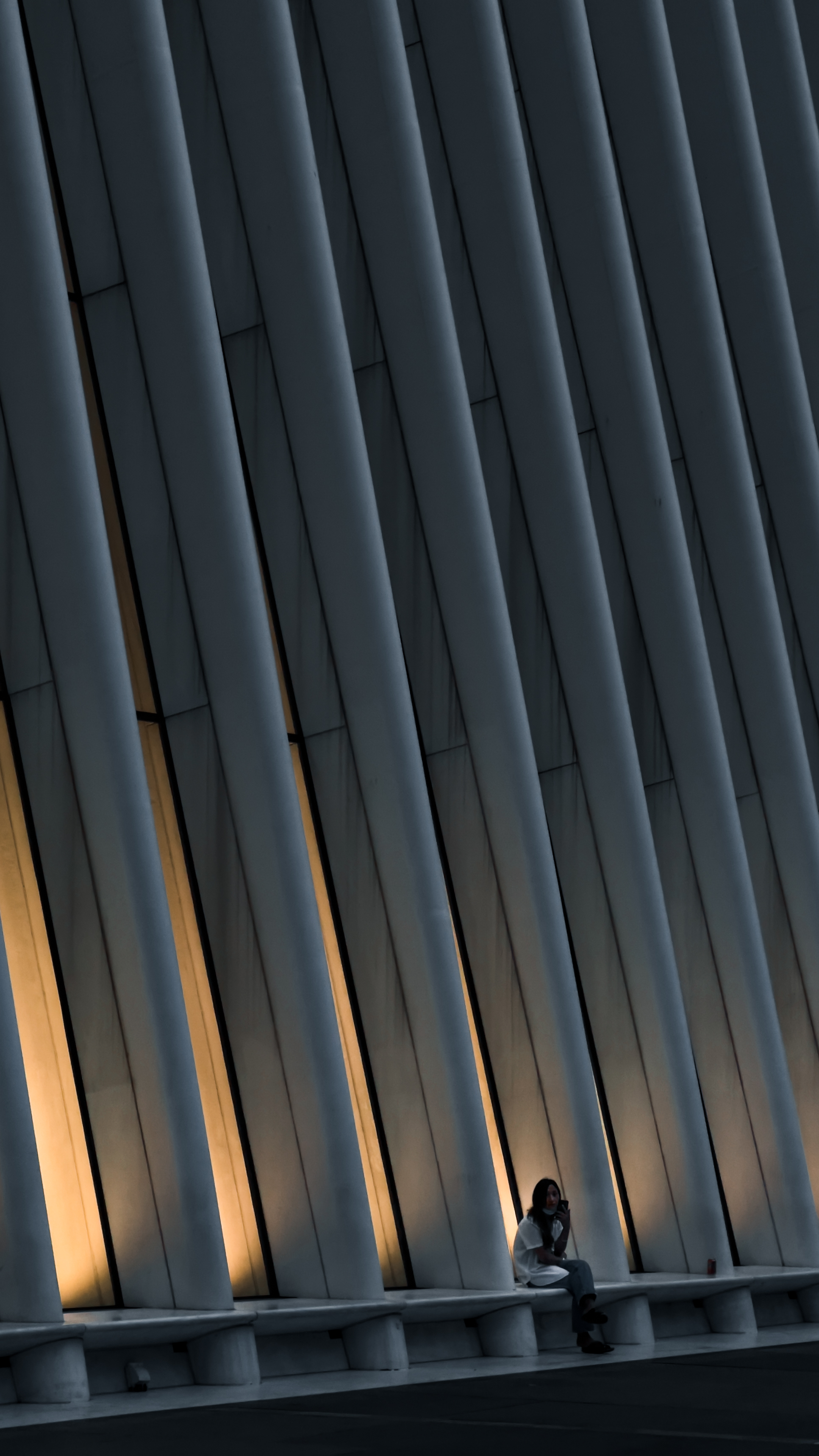 A person sits alone on a long bench against the dramatic, angled white beams
and soft yellow lighting of a modern architectural structure at
night.