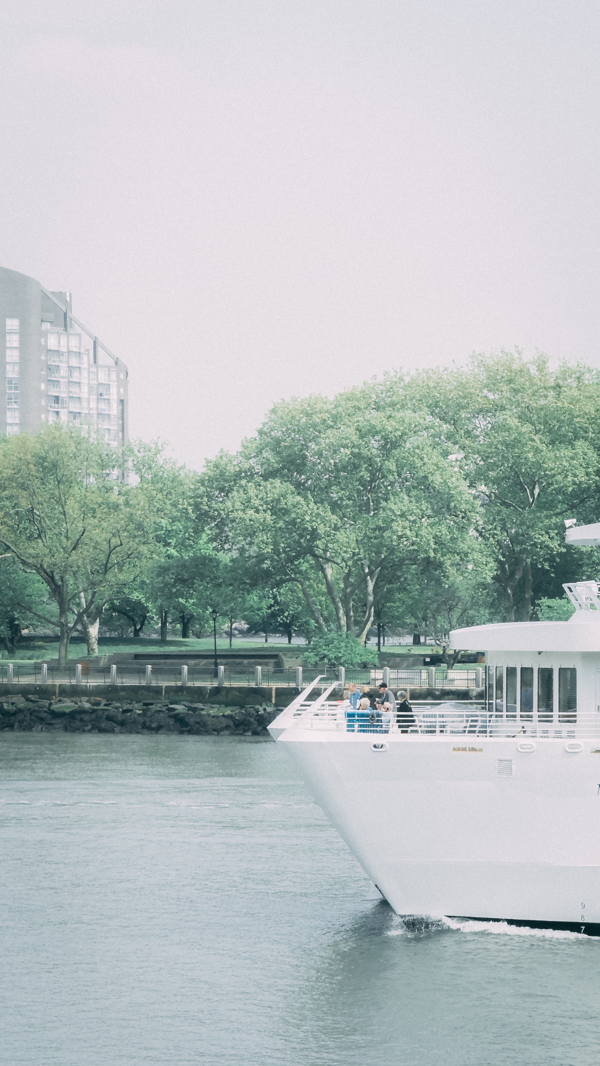 The bow of a white cruise ship with people gathered on the deck, passing by a
riverside park lined with green trees and city buildings in the
background.