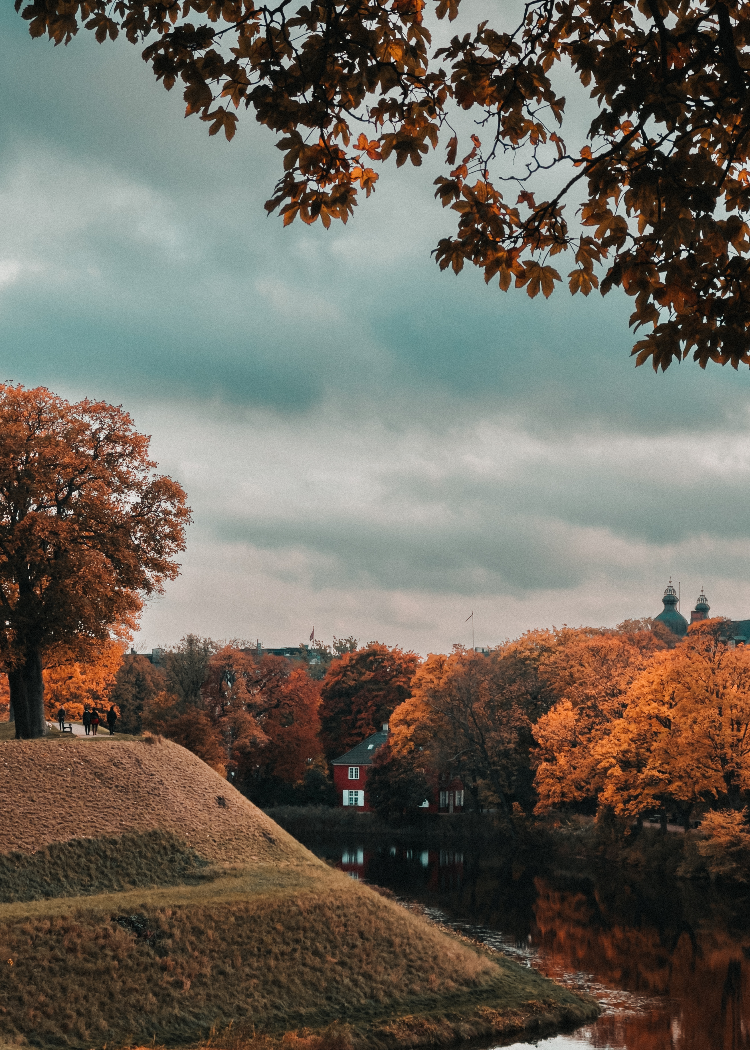 A grassy hill topped with autumn trees, people walking, and a calm moat
reflecting the fall colors, with dramatic clouds and a cityscape in the
distance.
