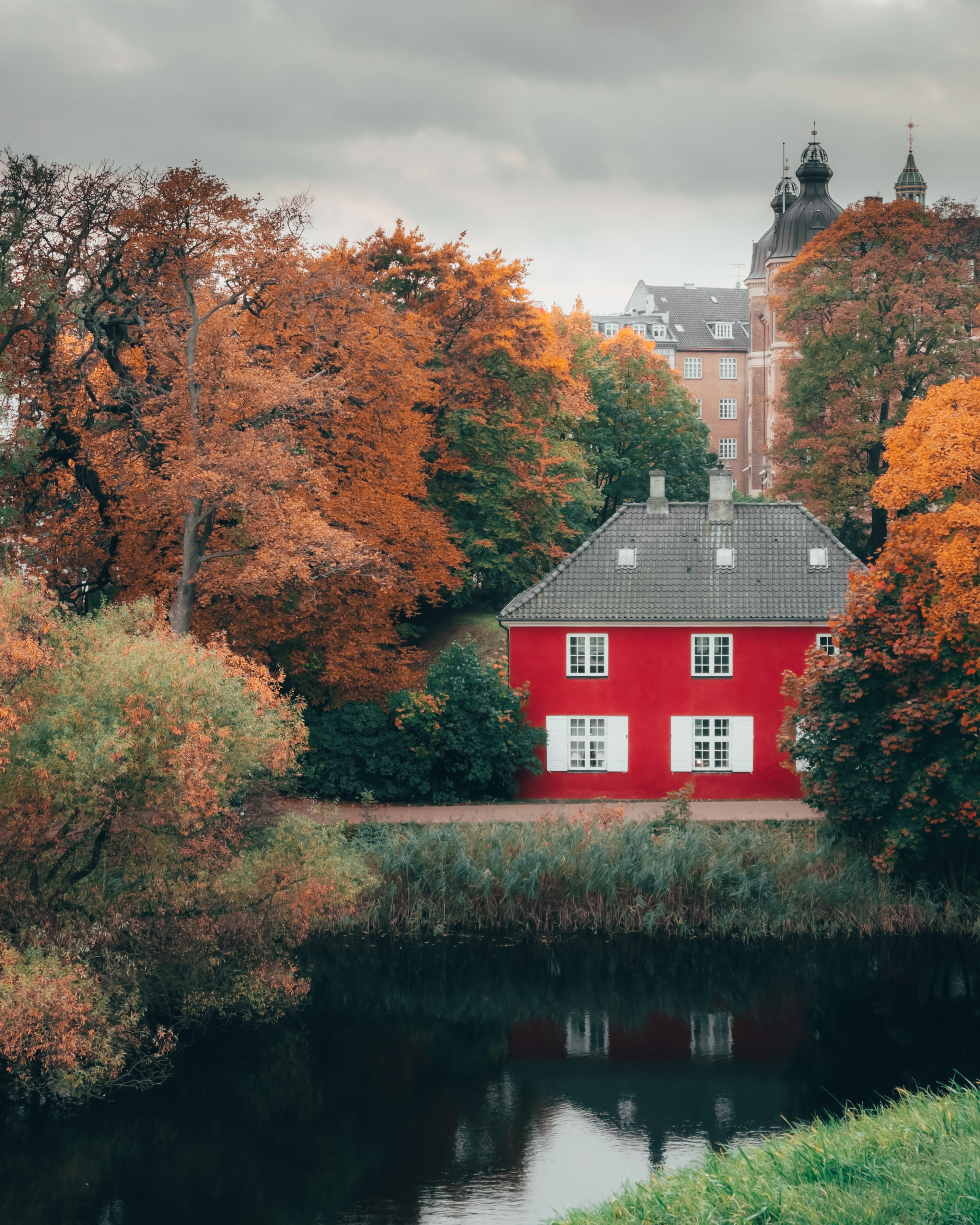 A bright red house with white shutters stands among colorful autumn trees
beside a reflective pond, with historic buildings in the background under an
overcast sky.