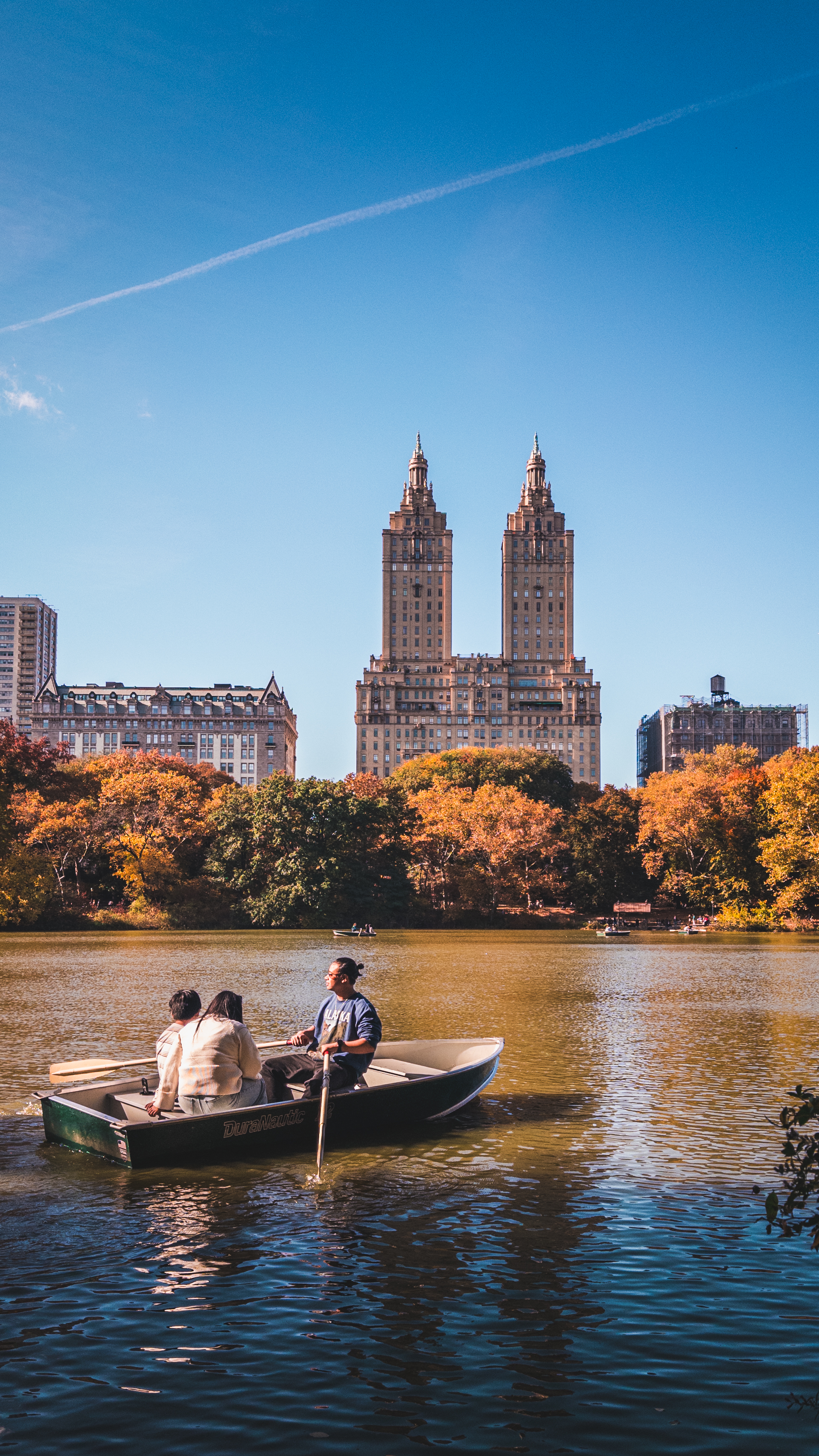 A man rowing two people in a small boat on a serene lake, with tall twin
towers and autumn trees in the background.