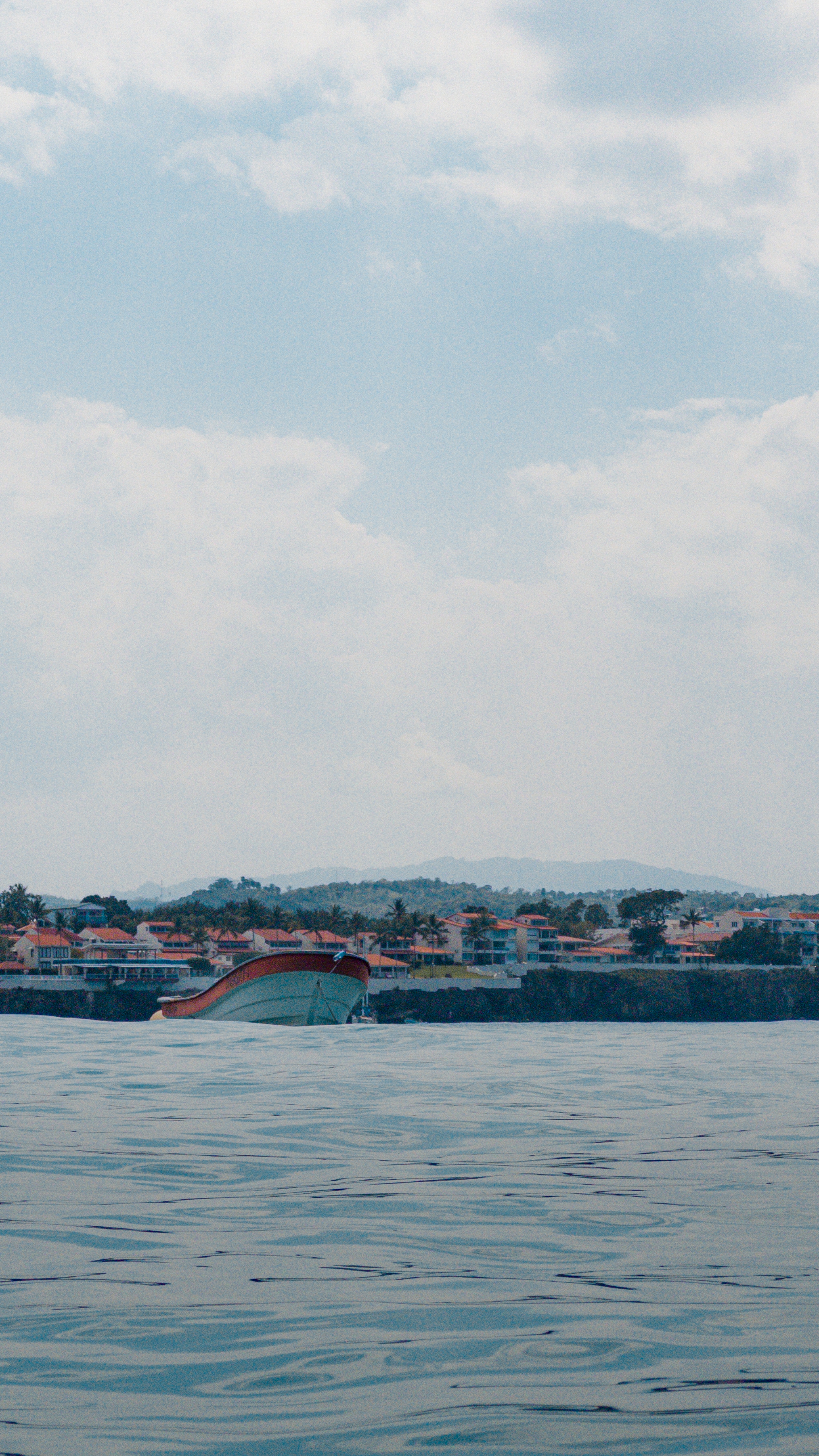 A small boat on calm ocean water near a distant shoreline with villas and palm
trees, under a partly cloudy sky.
