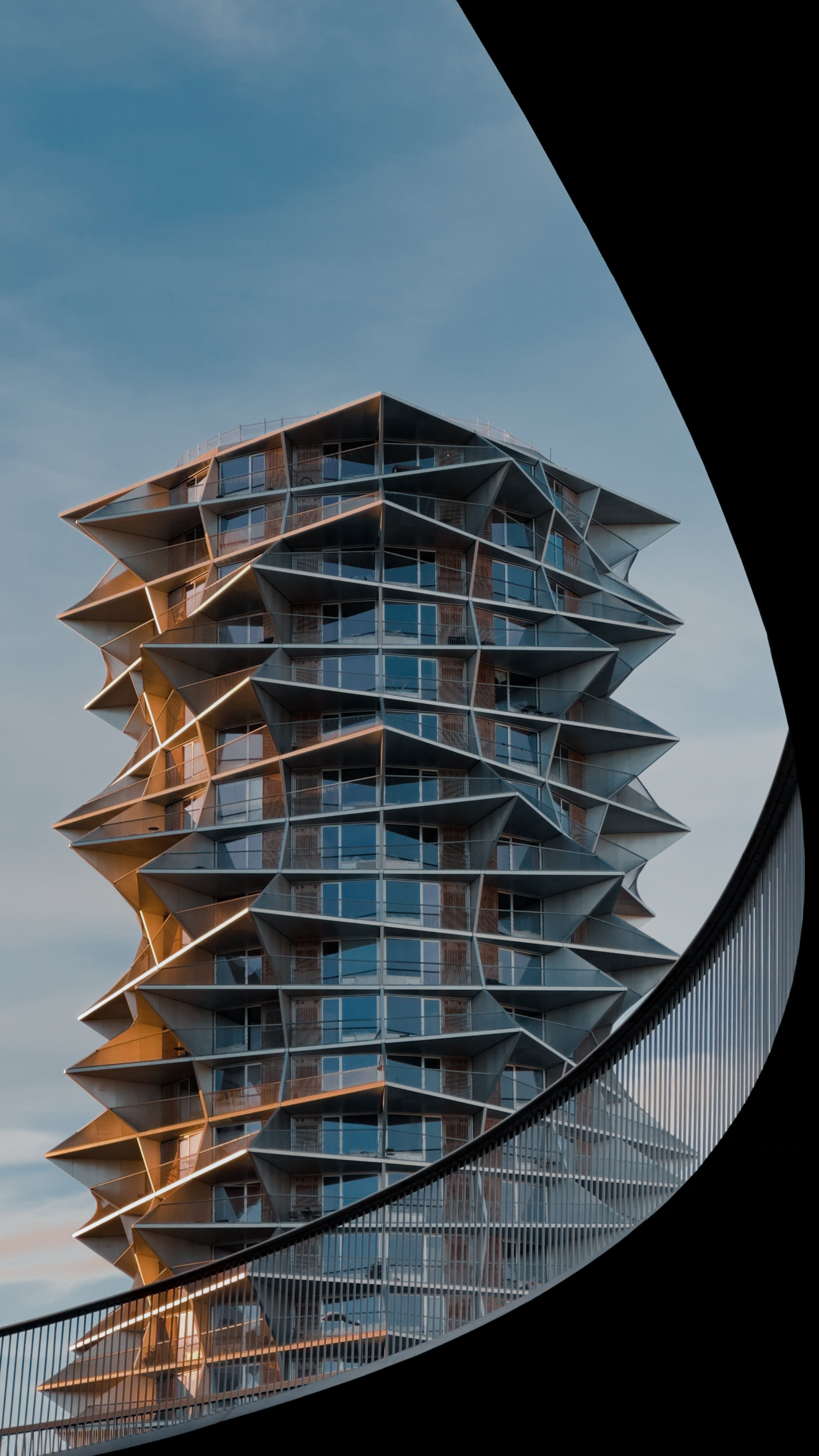 A modern architectural building with angular balconies, framed by a curving
bridge in the foreground and a blue sky in the
background.
