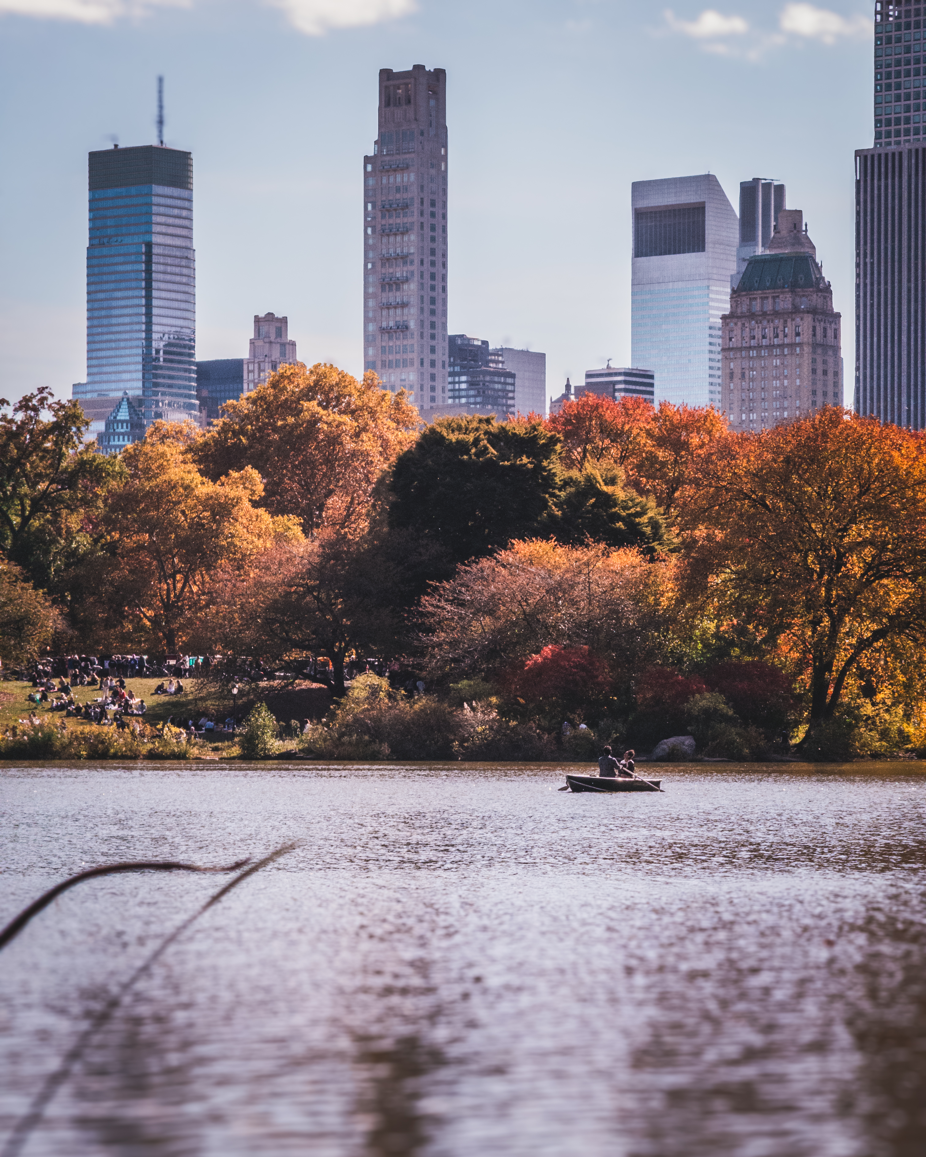 A boat with three people rowing on a calm lake, surrounded by autumn-colored
trees and skyscrapers in the distance.