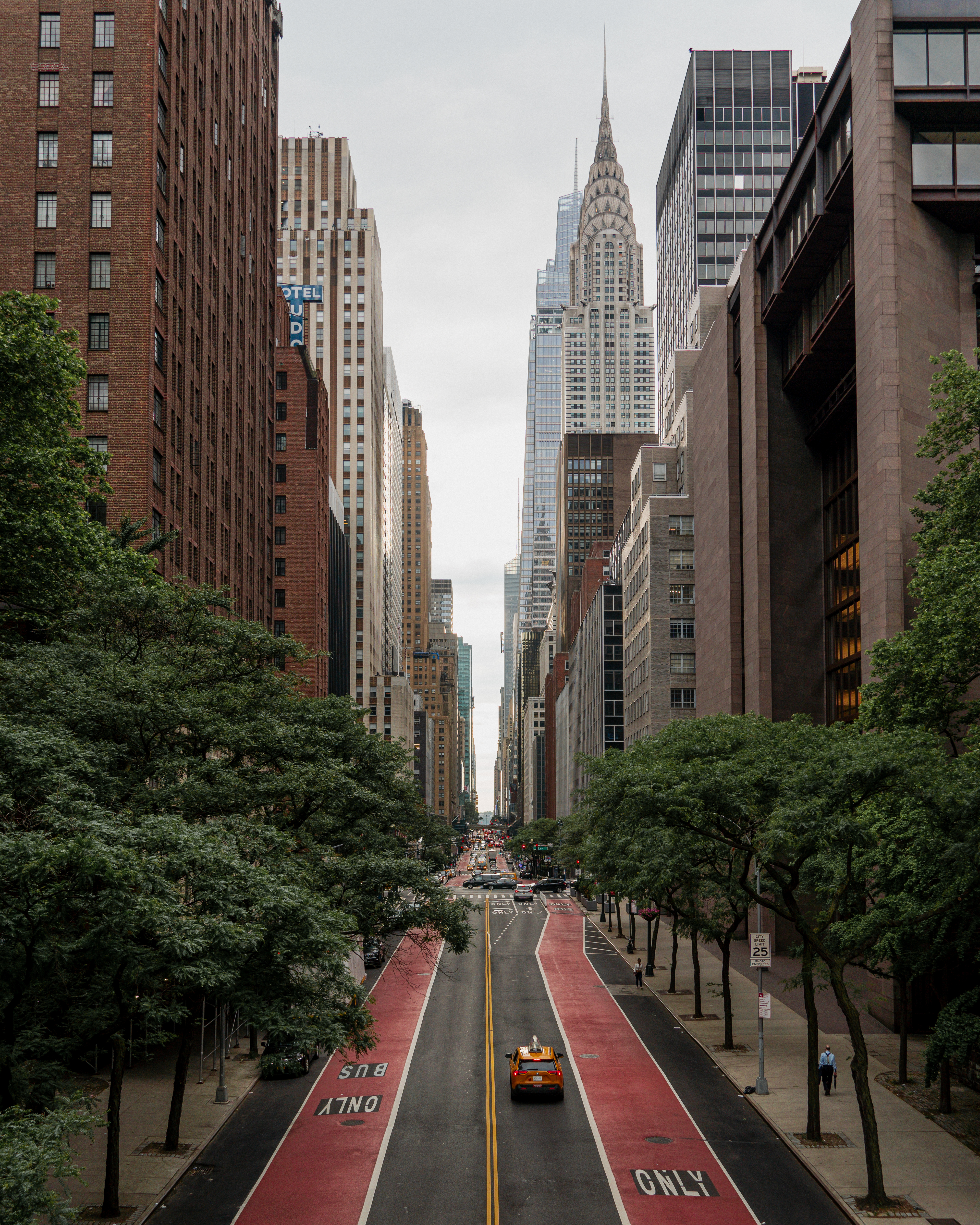 A view down a Manhattan street with tall skyscrapers on both sides, the
Chrysler Building in the background, trees lining the sidewalks, and a yellow
taxi driving along a red bus lane.
